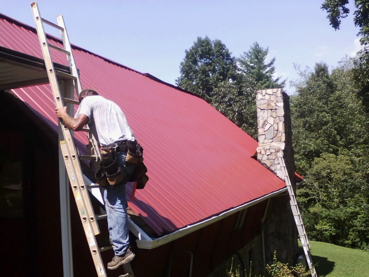 Roofer on a ladder beside a red metal roof and stone chimney — Soffit & Fascia Repair by Roofing Brothers in New Derry